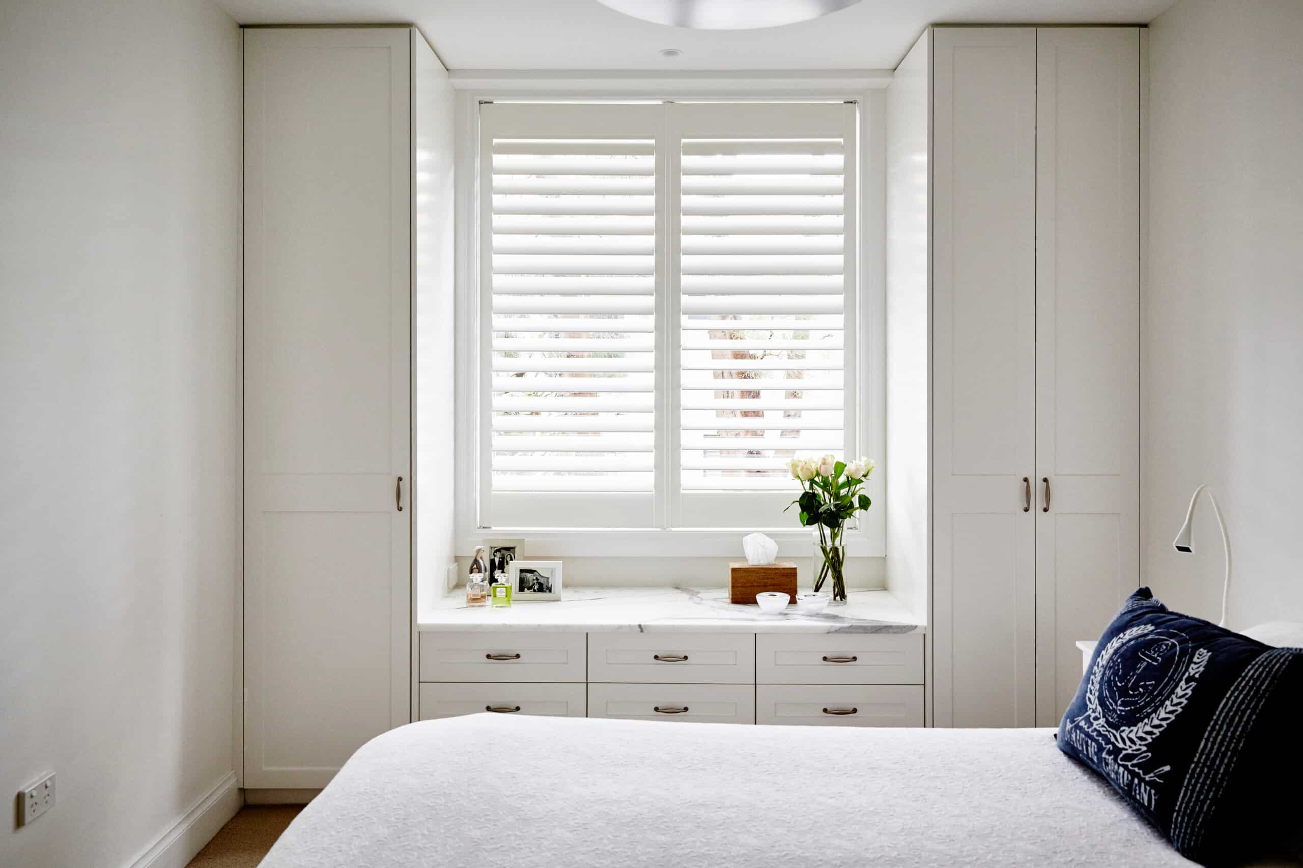 Interior photography of fresh and light bedroom with white bed, coastal style cushions, white built in cupboards and plantation shutters with decorative objects